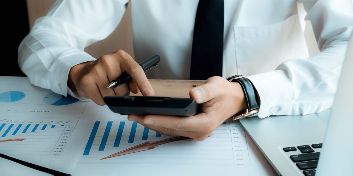 A man at a desk using a calculater with graphs and a computer in front of him-1