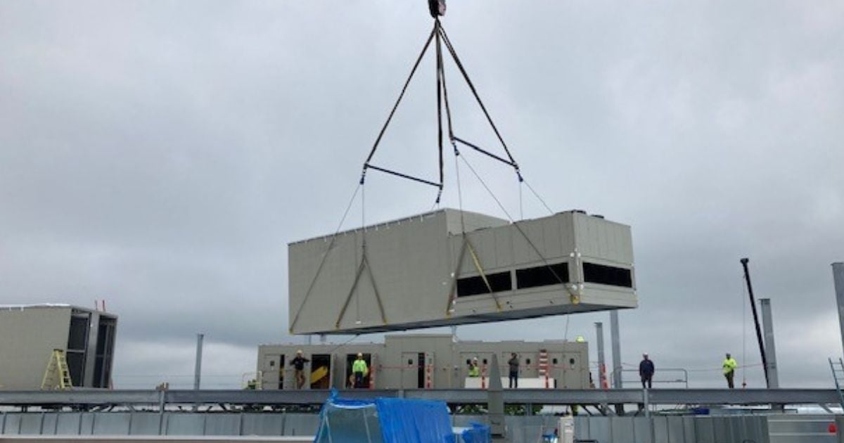 Large rooftop HVAC unit being lifted by crane onto a commercial building during overcast weather, with crew members guiding installation on site.