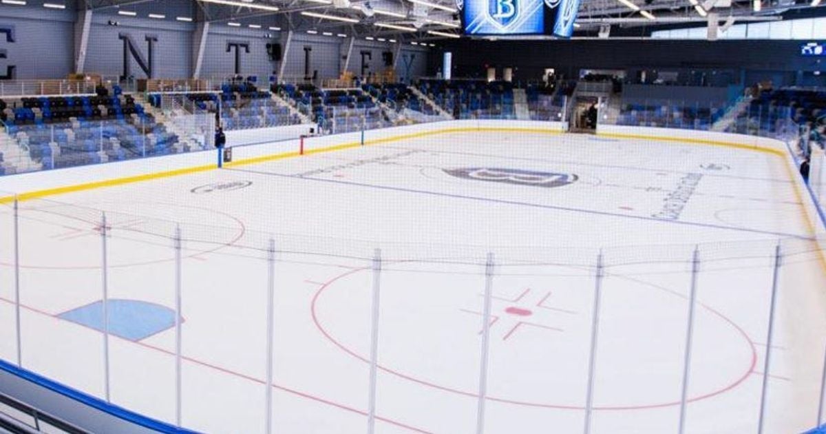 Empty indoor ice hockey arena with blue and gray seating, scoreboard overhead, and Bentley University logo at center ice.