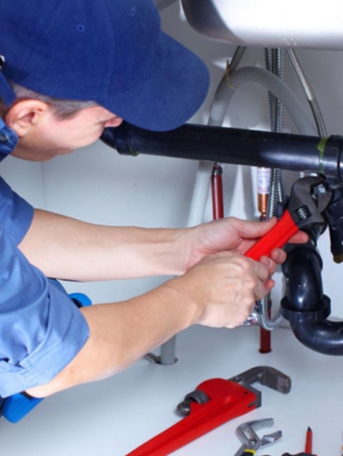 Plumber wearing a blue cap and shirt using a red pipe wrench to tighten black PVC pipes under a sink, with additional tools placed nearby.