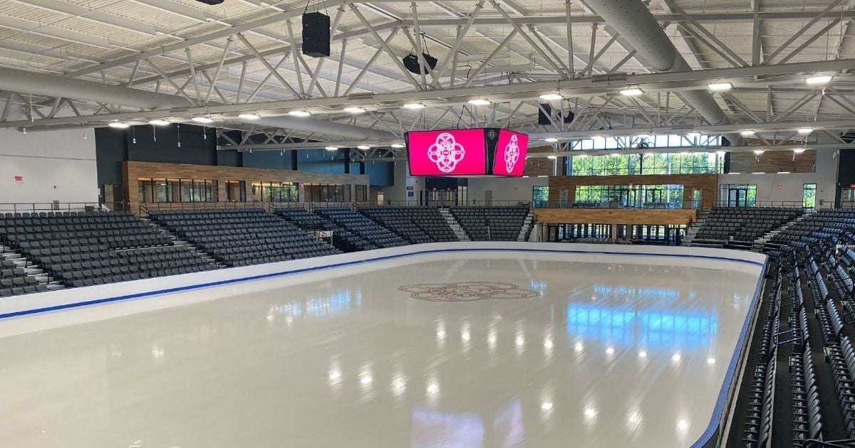 Modern indoor ice rink with polished surface, black seating, large digital scoreboard, and natural light streaming through floor-to-ceiling windows.