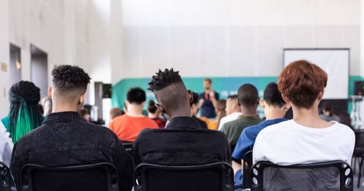 Students sitting in a school auditorium, listening to a speaker at the front of the room, with a projector screen and stage in the background.