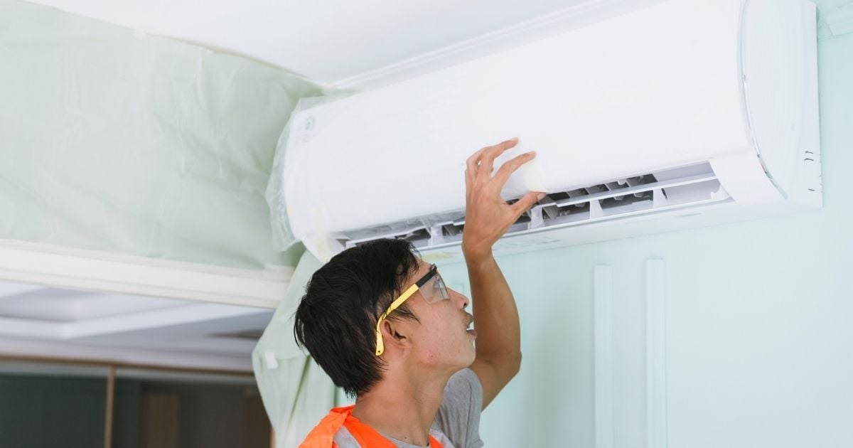 HVAC technician inspecting or servicing a wall-mounted mini-split air conditioning unit inside a building, wearing safety glasses and an orange safety vest.