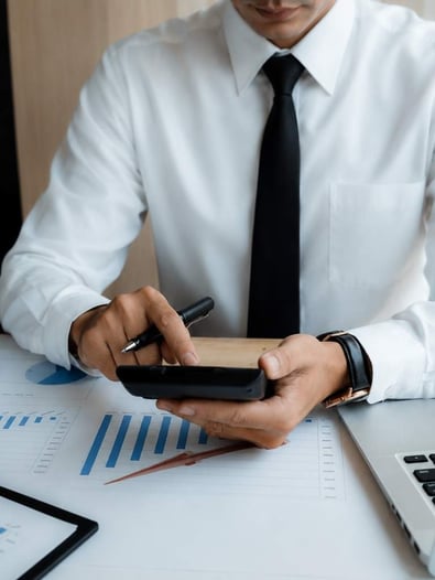 A man at a desk using a calculater with graphs and a computer in front of him